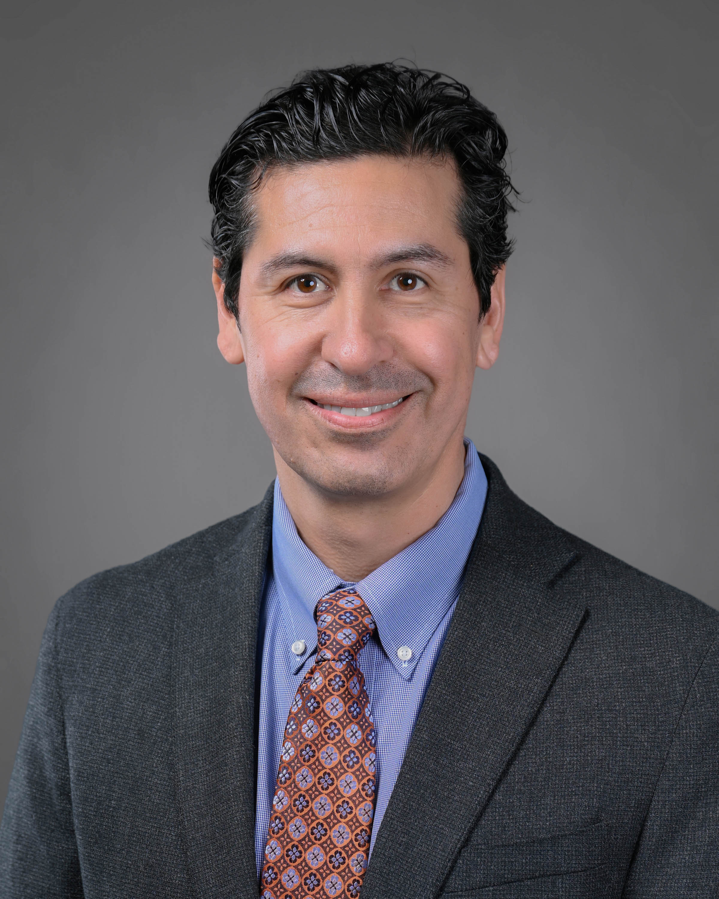 Kind man smiling with dark hair in a dark gray jacket with blue shirt and orange patterned tie.
